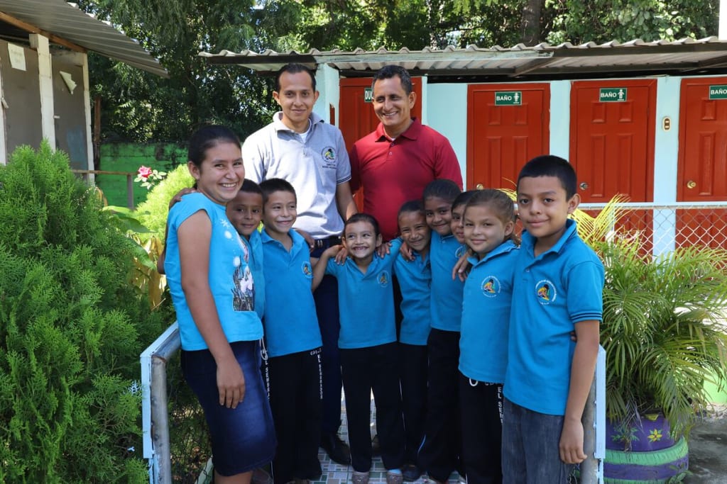 A group of children in blue shirts stand with a man in red and another man in white